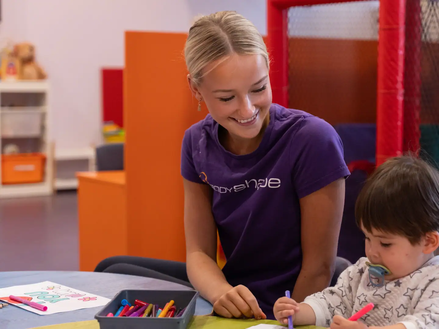 A Kids Club staff member sitting with a young child at the activity table inside Body Shape Warringah Mall